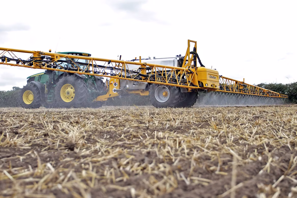 A tractor with a wide boom sprayer crossing a harvested field, seen from ground level.