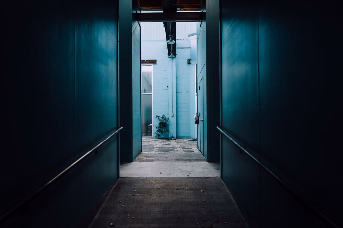 A narrow passage between dark teal walls opening onto a bright courtyard with a small plant.